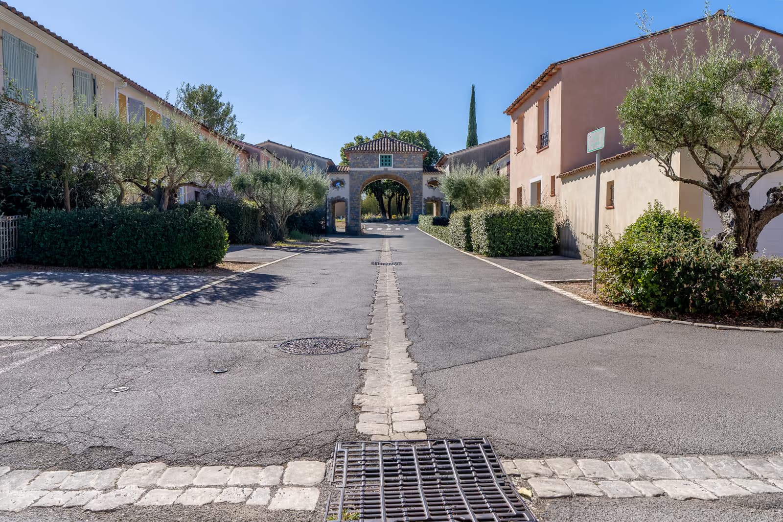 Porche en pierre voûté ouvrant sur une allée plantée d'oliviers au centre du village de Saint-Clément-de-Rivière