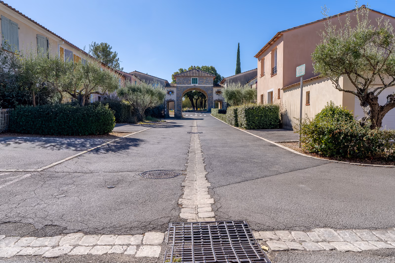 Porche en pierre voûté ouvrant sur une allée plantée d'oliviers au centre du village de Saint-Clément-de-Rivière