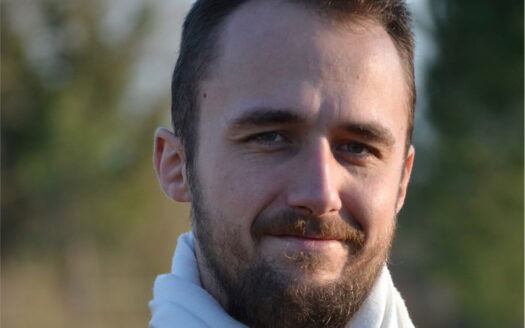 Homme avec barbe courte et écharpe blanche en extérieur avec arrière-plan flou d'arbres.