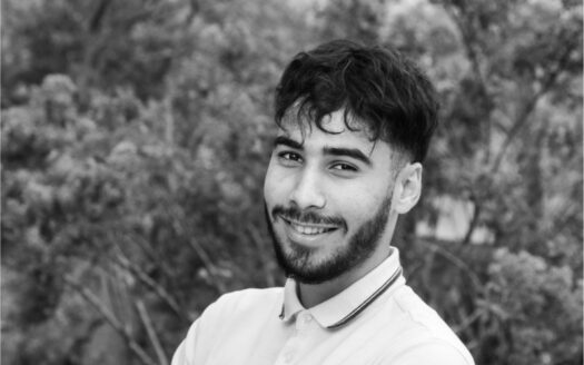 Homme souriant en polo blanc Boss devant un fond flou d’arbres.