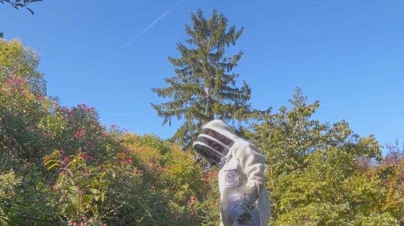 Personne en combinaison d'apiculteur debout près d'un pulvérisateur en plein air sous un ciel bleu.