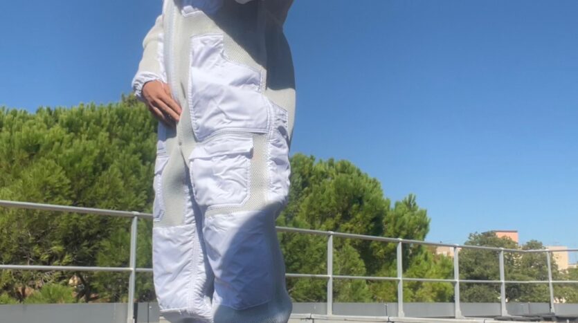 Un homme en combinaison apicole blanche avec cagoule sur un sol de gravier, ciel bleu et arbres.