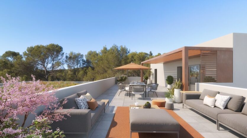 Terrasse avec canapés gris, table à manger sous parasol, plantes et arbres sous ciel bleu.