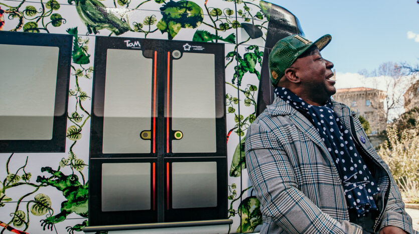 Homme avec casquette verte et écharpe à pois assis près d'un tram décoré de motifs végétaux.