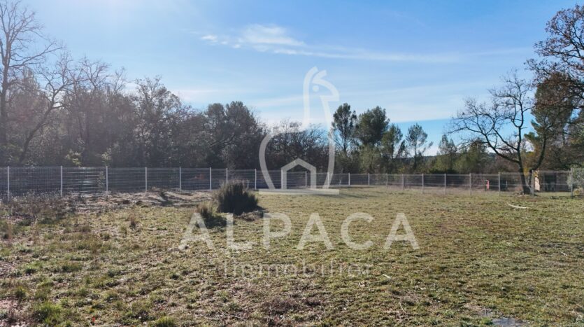 Terrain plat enherbé clôturé, arbres clairsemés en arrière-plan sous ciel bleu partiellement nuageux.