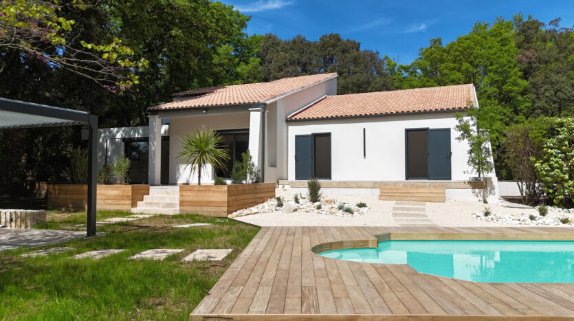 Maison moderne avec terrasse en bois, piscine, jardin gazonné et cailloux blancs, entourée d'arbres.