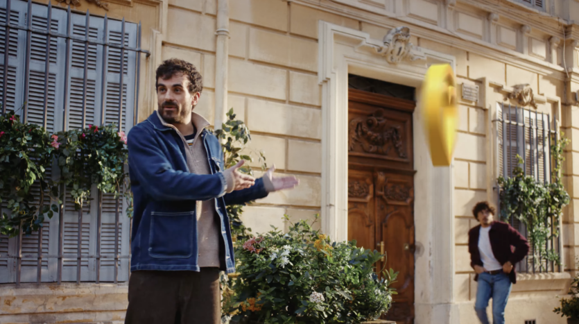 Deux hommes dans une rue avec un bâtiment orné et un objet jaune suspendu entre eux.