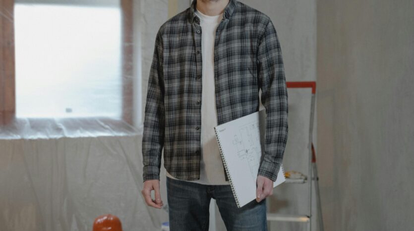 Homme avec casque blanc et lunettes de protection tenant plans dans pièce en rénovation avec outils.